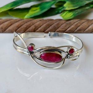May include: A silver cuff bracelet with three oval-shaped, deep red gemstones. The bracelet has an open design with a swirling pattern around the central stone. The gemstones are set in a polished silver metal.