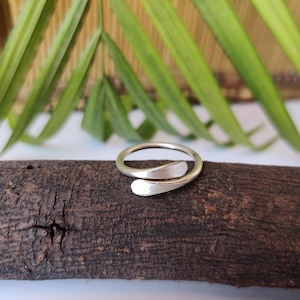 Può includere: Un anello color argento dal design aperto. L'anello presenta due forme curve e allungate che si sovrappongono. L'anello è esposto su un pezzo di legno marrone scuro, con foglie verdi sullo sfondo.