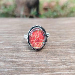 Puede incluir: Anillo de plata ovalado con una piedra roja e iridiscente. La piedra está engastada en un borde trenzado. El anillo se exhibe sobre una superficie de madera, mostrando su diseño y color.