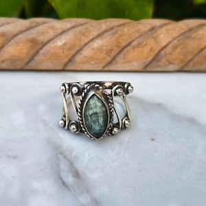 Puede incluir: Un anillo de plata con una piedra preciosa verde ovalada en el centro. El anillo tiene un diseño ornamentado con remolinos decorativos y pequeñas cuentas de plata. La piedra preciosa está engastada en un borde en forma de cuerda. El anillo se muestra sobre una superficie de mármol blanco y gris.