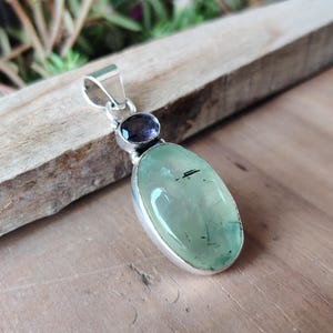 May include: A silver pendant featuring a large, oval-shaped, light green gemstone with black inclusions, paired with a smaller, round, dark purple stone. The pendant has a silver bail for a chain.