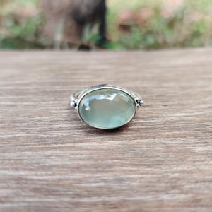 May include: A silver ring featuring a large, oval, pale green gemstone. The ring has a simple band and small decorative elements either side of the gemstone. The ring is set against a wooden surface.