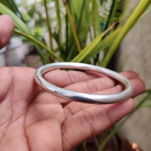 Pode incluir: Uma pulseira de prata com um design liso e arredondado. A pulseira é segurada em uma mão, com folhagem verde ao fundo. A pulseira é uma peça de joalheria simples e elegante.