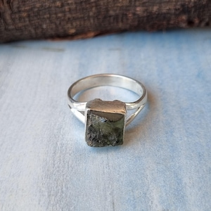 Puede incluir: Un anillo de plata con una piedra preciosa cuadrada de color verde oscuro engastada en el centro. El anillo tiene un diseño simple y elegante.