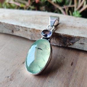 May include: A silver pendant featuring a large, oval, pale green gemstone with a dark inclusion. Above it is a smaller, oval, purple gemstone. The pendant has a silver bail for a chain. The jewelry is displayed on a wooden surface.