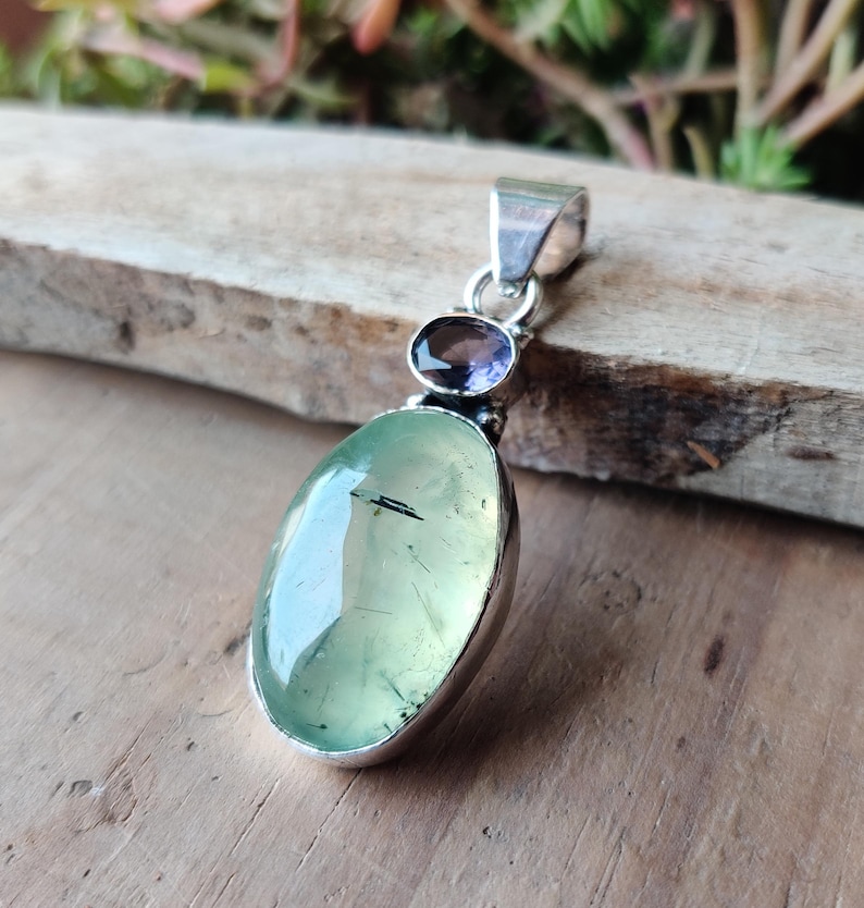 May include: A silver pendant featuring a large, oval, pale green gemstone with a dark inclusion. Above it is a smaller, oval, purple gemstone. The pendant has a silver bail for a chain. The jewelry is displayed on a wooden surface.