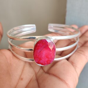May include: A silver cuff bracelet with a large, oval-cut ruby gemstone. The bracelet has three parallel bands. The ruby is a vibrant red, and the silver has a polished finish. The bracelet is open-ended.
