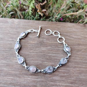 Puede incluir: Una pulsera de plata con piedras de cuarzo rosa ovaladas. La pulsera tiene un cierre de palanca y está compuesta por eslabones interconectados. Las piedras de cuarzo rosa están engastadas en biseles de plata ornamentados, creando un diseño delicado y elegante.