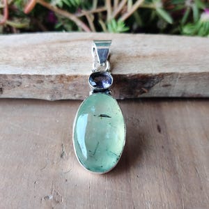 May include: A silver pendant featuring an oval, light green gemstone with black inclusions, topped by a smaller, round, purple gemstone. The pendant has a silver bail for a chain. The jewelry is displayed on a wooden surface.