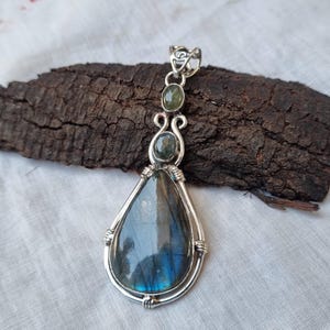 May include: A silver pendant with a large teardrop-shaped labradorite gemstone and two smaller green gemstones. The pendant is set in a silver wire frame.