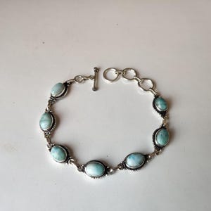 May include: A silver bracelet featuring oval-shaped, light blue gemstones. The bracelet has a toggle clasp and is composed of interconnected links. The gemstones are set in a decorative silver setting.