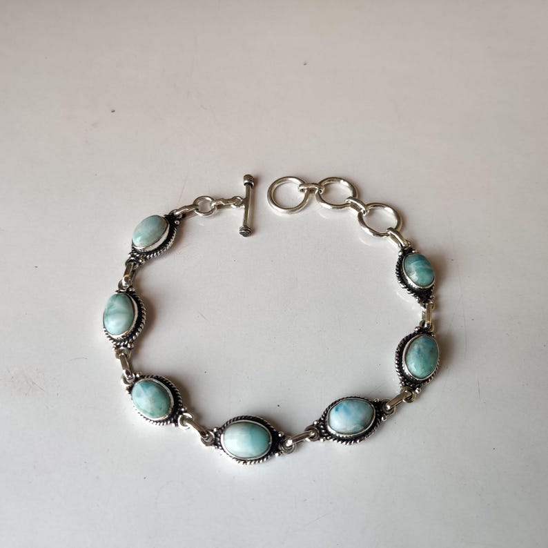May include: A silver bracelet featuring oval-shaped, light blue gemstones. The bracelet has a toggle clasp and is composed of interconnected links. The gemstones are set in a decorative silver setting.