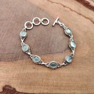 May include: A silver bracelet featuring oval, pale blue gemstones. The bracelet has a toggle clasp and is set on a wooden surface. The gemstones are framed by a decorative silver border.