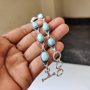 May include: A silver bracelet featuring oval, light blue gemstones. The gemstones are set in ornate silver settings and connected by silver links. The bracelet has a toggle clasp for closure. The bracelet is approximately 7 inches long.