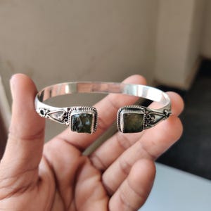 May include: A silver cuff bracelet featuring two square, green-toned stones. The bracelet has decorative detailing near the stones. The bracelet is held in a hand.