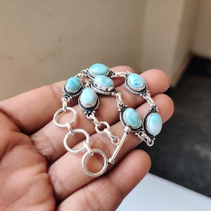 May include: A silver bracelet featuring oval-shaped, light blue gemstones. The gemstones are set in ornate silver settings and connected by silver links. The bracelet has a toggle clasp closure. The bracelet is displayed against a neutral background.