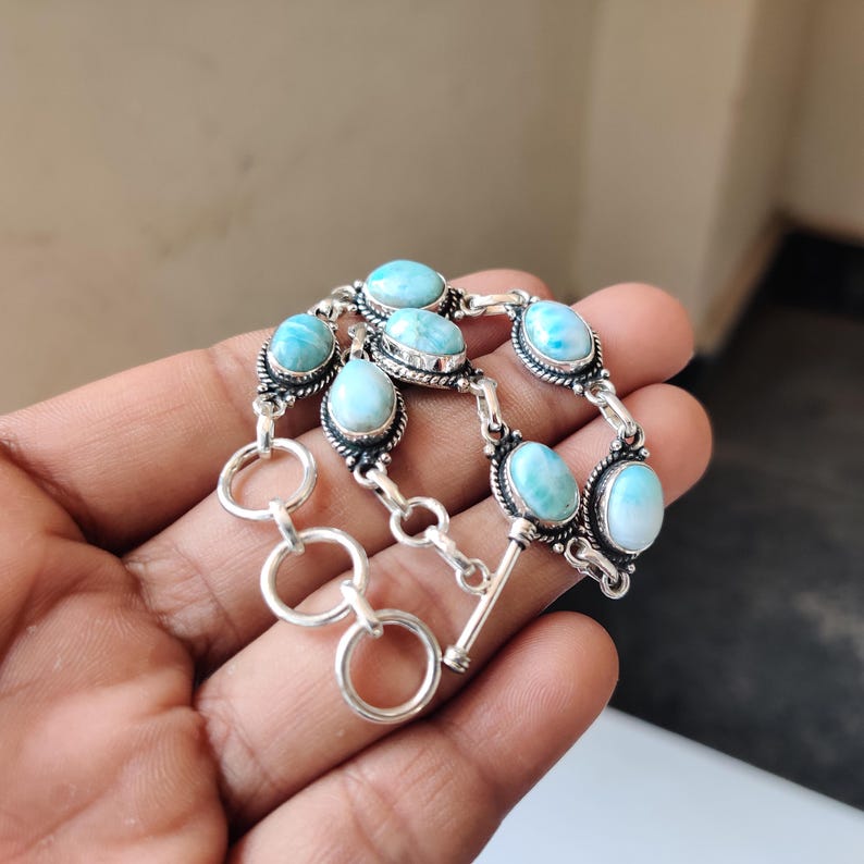 May include: A silver bracelet featuring oval-shaped, light blue gemstones. The gemstones are set in ornate silver settings and connected by silver links. The bracelet has a toggle clasp closure. The bracelet is displayed against a neutral background.