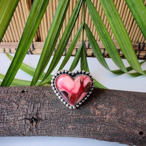 May include: A heart-shaped ring with a pink and red stone set in a silver beaded frame. The ring is displayed on a piece of wood, with green leaves in the background. The ring's stone has a unique, marbled appearance.
