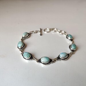 May include: A silver bracelet featuring oval, light blue gemstones. The gemstones are set in ornate silver settings and connected by silver links. The bracelet has a toggle clasp.