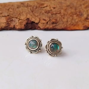 Könnte beinhalten: Ein Paar silberne Ohrstecker mit floralem Design. Jeder Ohrring hat einen runden, irisierenden, teal-farbenen Stein, der in einen dekorativen Silberrahmen gefasst ist. Die Ohrringe werden auf einer weißen Oberfläche präsentiert.
