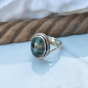 Puede incluir: Un anillo de plata con una piedra de ágata musgosa ovalada. La piedra muestra patrones verdes y blancos, engastada en un bisel decorativo. La banda del anillo tiene un diseño dividido.