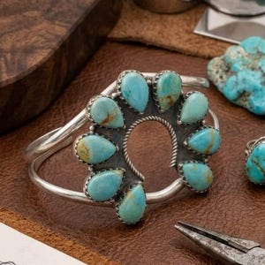 Op de afbeelding: Een zilveren manchetarmband met een cirkelvormige opstelling van negen druppelvormige turkoois stenen. De stenen zijn gezet in zilveren zettingen met een gedraaide draadrand rondom de centrale opening. De armband heeft een gladde band.
