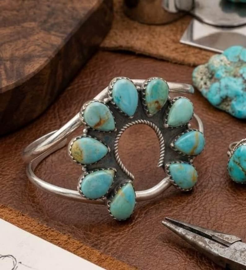 Op de afbeelding: Een zilveren manchetarmband met een cirkelvormige opstelling van negen druppelvormige turkoois stenen. De stenen zijn gezet in zilveren zettingen met een gedraaide draadrand rondom de centrale opening. De armband heeft een gladde band.