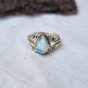May include: A silver ring featuring an oval opal gemstone. The ring has an intricate, filigree design around the gemstone and on the band. The opal displays a play of colours, including blue, green, and yellow. The ring is set against a white background.