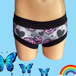May include: Underwear featuring a gray background with a pattern of black hearts, small stars, and pink leopard-print hearts. The garment has a wide black waistband and black leg openings, providing a contrasting border to the patterned center panel.