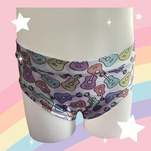 May include: Underwear featuring a repeating pattern of acoustic guitars in pastel shades of purple, blue, green, and pink on a white background. The garment has a wide waistband and a fitted cut, displaying a colorful musical instrument motif throughout.