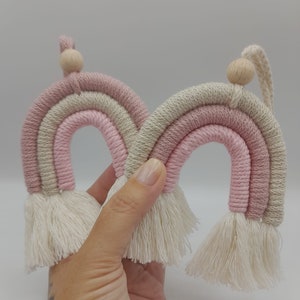 Könnte beinhalten: Zwei rosa und beigefarbene Makramee-Regenbögen mit weißen Quasten, die an Holzperlen hängen.