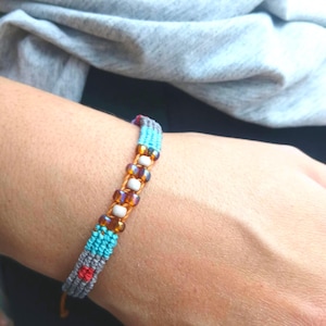 Könnte beinhalten: Ein handgefertigtes Makramee-Armband mit einem gewebten Design in Grau, Türkis und Rot. Das Armband ist mit bernsteinfarbenen und weißen Perlen verziert. Das Armband wird am Handgelenk getragen.
