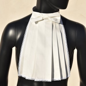 Puede incluir: Un babero blanco plisado con ribete de encaje y una pajarita. El babero se lleva sobre un maniquí negro.