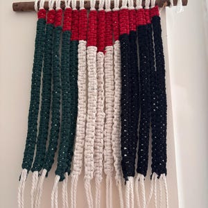 May include: A wall hanging made of woven string in the colours of the United Arab Emirates flag: green, white, red, and black. The hanging is attached to a wooden dowel.