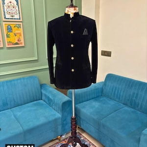 Puede incluir: Una chaqueta de terciopelo negro con cuello mandarín y botones dorados se exhibe en un maniquí. Un pañuelo de bolsillo negro está en el bolsillo del pecho. La chaqueta forma parte de una colección de kurtas personalizadas, como indica el texto en la parte inferior de la imagen.