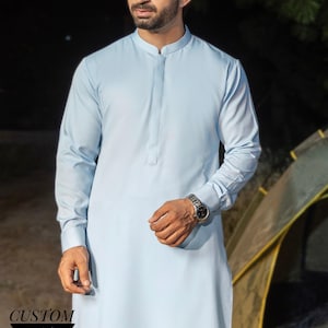Puede incluir: Un kurta azul claro con cuello mandarín y mangas largas. La imagen muestra a un hombre usando el kurta, con un reloj en la muñeca. El texto "CUSTOM KURTAS" se muestra en una pancarta negra en la parte inferior de la imagen.