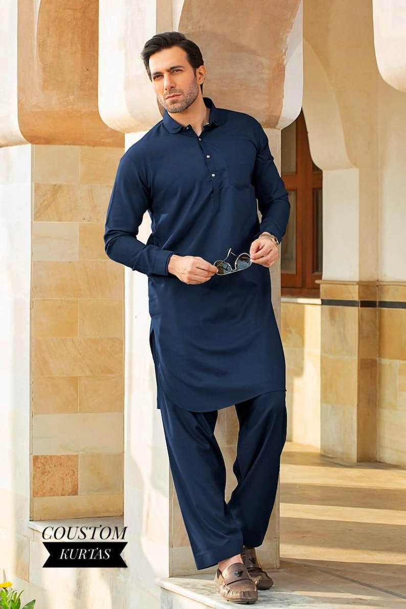 May include: A navy blue long-sleeved kurta with a button-up collar and a pocket on the left side. The kurta is paired with matching navy blue pants. The man is wearing brown loafers and holding a pair of sunglasses.