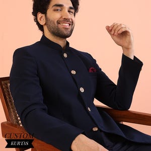 以下が含まれることがあります： 金色のボタンとマオカラーが付いたネイビーブルーのジャケット。胸ポケットには赤いポケットチーフが見えます。ジャケットはスーツの一部です。「CUSTOM KURTAS」の文字が黒いバナーに印刷されています。