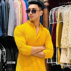 Pu&ograve; includere: Un kurta giallo brillante a maniche lunghe con un intricato motivo ricamato tono su tono, colletto alla coreana e abbottonatura. Il capo &egrave; esposto in un negozio con il testo CUSTOM KURTAS visibile nell'angolo in basso a sinistra.