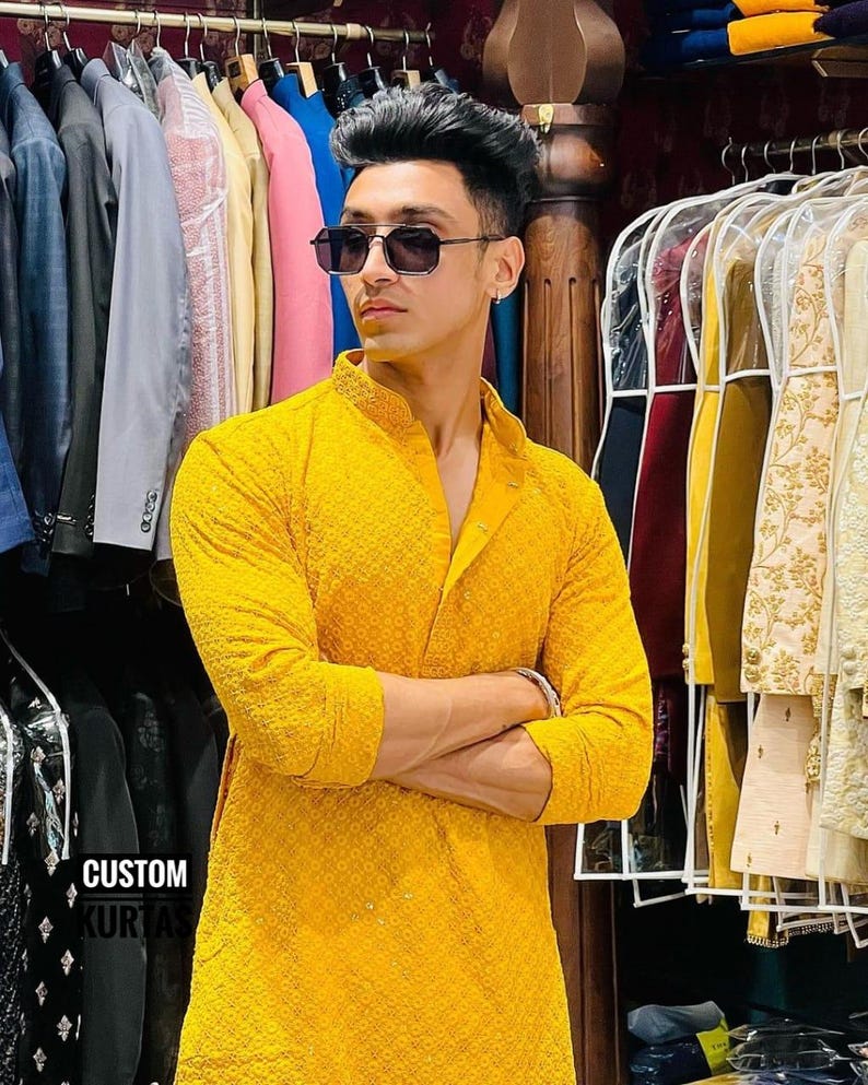 Pu&ograve; includere: Un kurta giallo brillante a maniche lunghe con un intricato motivo ricamato tono su tono, colletto alla coreana e abbottonatura. Il capo &egrave; esposto in un negozio con il testo CUSTOM KURTAS visibile nell'angolo in basso a sinistra.