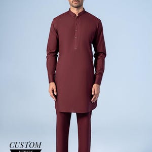 Puede incluir: Un traje kurta burdeos con cuello mandarín y mangas largas. Los pantalones a juego completan el conjunto. El texto "CUSTOM KURTAS" se muestra en la parte inferior de la imagen.