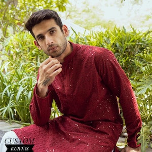 Puede incluir: Un kurta burdeos con detalles intrincados y sutiles adornos. La prenda de manga larga presenta un cuello mandarín y se combina con pantalones blancos. La imagen incluye el texto "CUSTOM KURTAS".