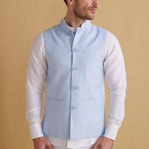 Pode incluir: Um colete de linho azul claro com gola mandarim e seis botões. O colete tem dois bolsos e é usado sobre uma camisa branca.