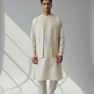May include: Cream-colored kurta set with a matching vest. The long-sleeved shirt has a mandarin collar and pleated front. The pants are straight-legged. The outfit is paired with brown sandals. The text "CUSTOM KURTAS" is at the bottom.