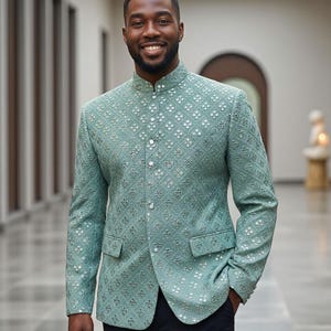Può includere: Giacca verde acqua con collo alla coreana e un sottile motivo floreale. La giacca ha una chiusura con bottoni sul davanti e due tasche anteriori. La persona indossa pantaloni neri.