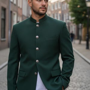 Puede incluir: Un traje verde oscuro con cuello mao, botones plateados y un bolsillo en el pecho. El traje se lleva con una camiseta blanca. El fondo muestra una calle adoquinada y edificios.
