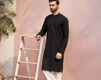 May include: A black long-sleeved kurta with a button-up collar and a white button. The kurta is worn with white pants and black shoes. The model is standing in front of a pink wall with a wooden ladder.