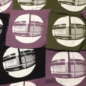 May include: A selection of fabric patches in shades of black, purple, and olive green. Each patch displays a white graphic of a building facade within a white circle. The patches are arranged on a wooden surface.