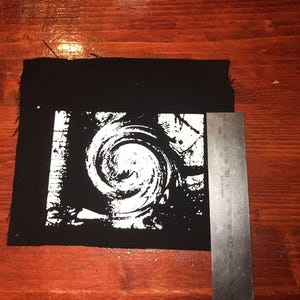 Puede incluir: Parche cuadrado negro con un dise&ntilde;o de espiral blanco. Una regla met&aacute;lica est&aacute; al lado del parche. El parche mide aproximadamente 10 cm de ancho.