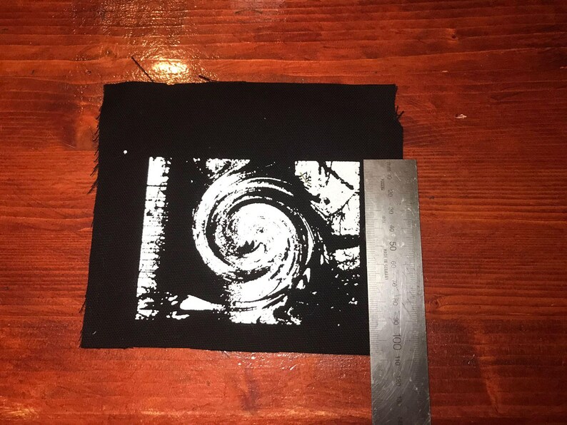 Puede incluir: Parche cuadrado negro con un dise&ntilde;o de espiral blanco. Una regla met&aacute;lica est&aacute; al lado del parche. El parche mide aproximadamente 10 cm de ancho.